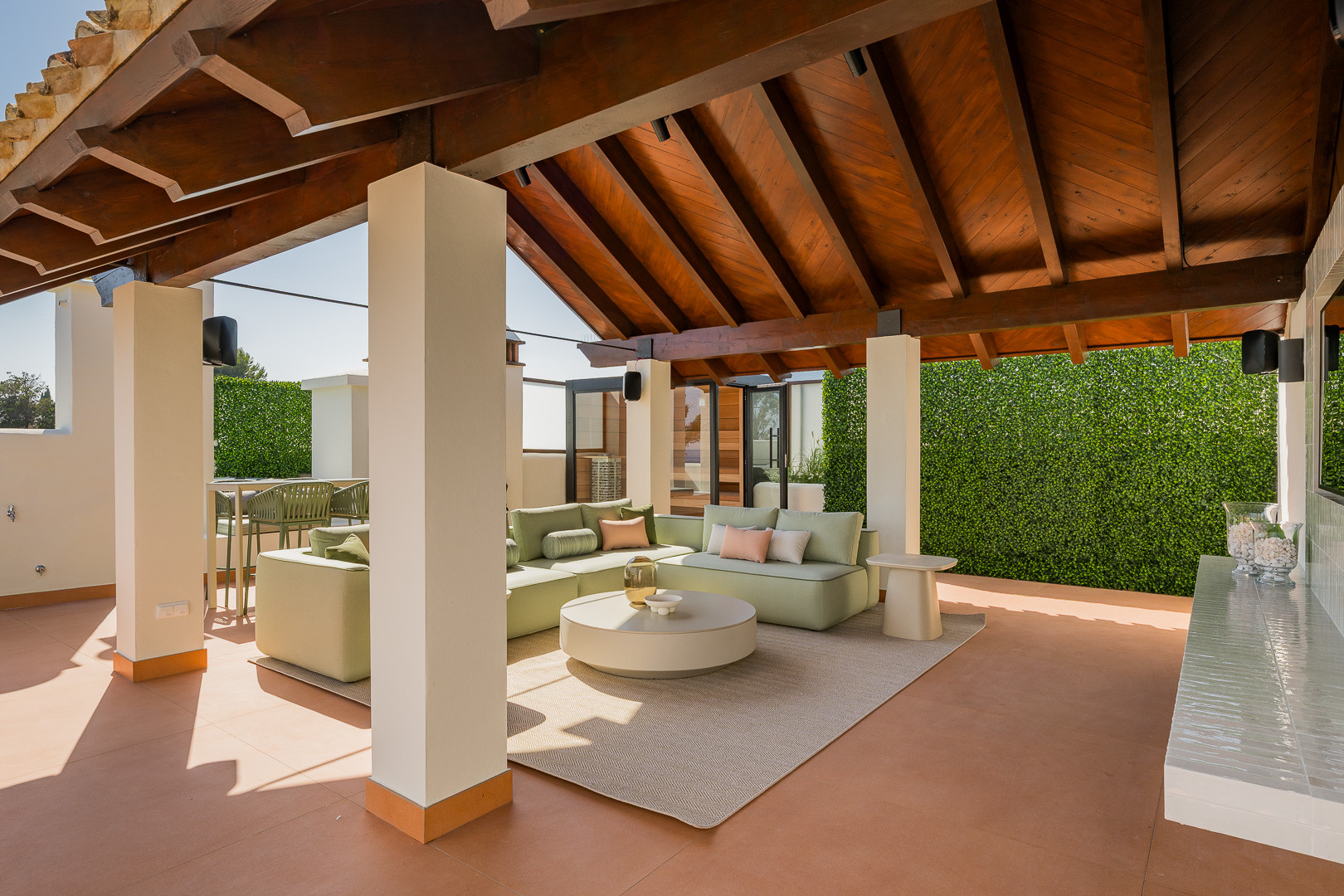 Covered outdoor lounge area with green sofas, coffee table, and exposed wooden beams.