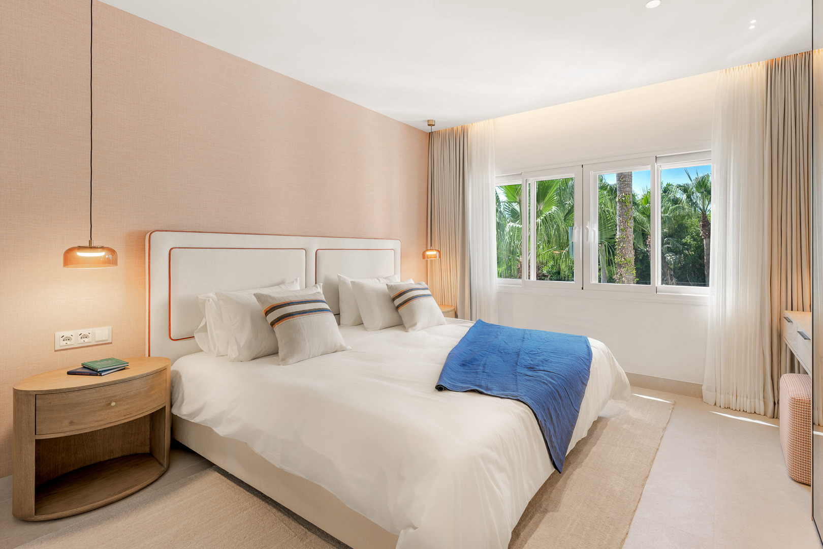 Bedroom with a large bed, textured peach wall, and palm trees visible through the window.