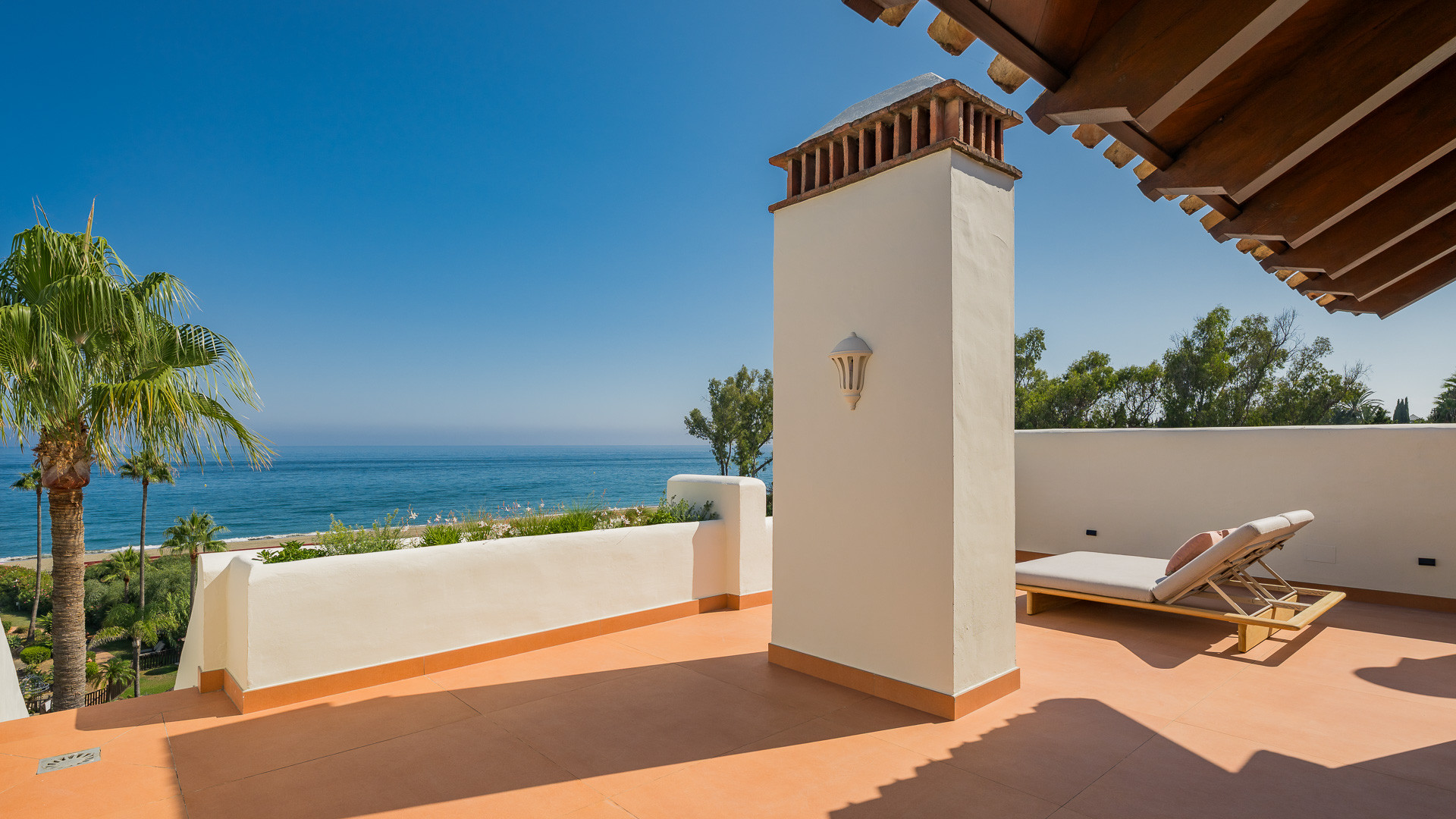 Outdoor terrace with orange floor, chimney, sun lounger, and view of the ocean and palm trees.