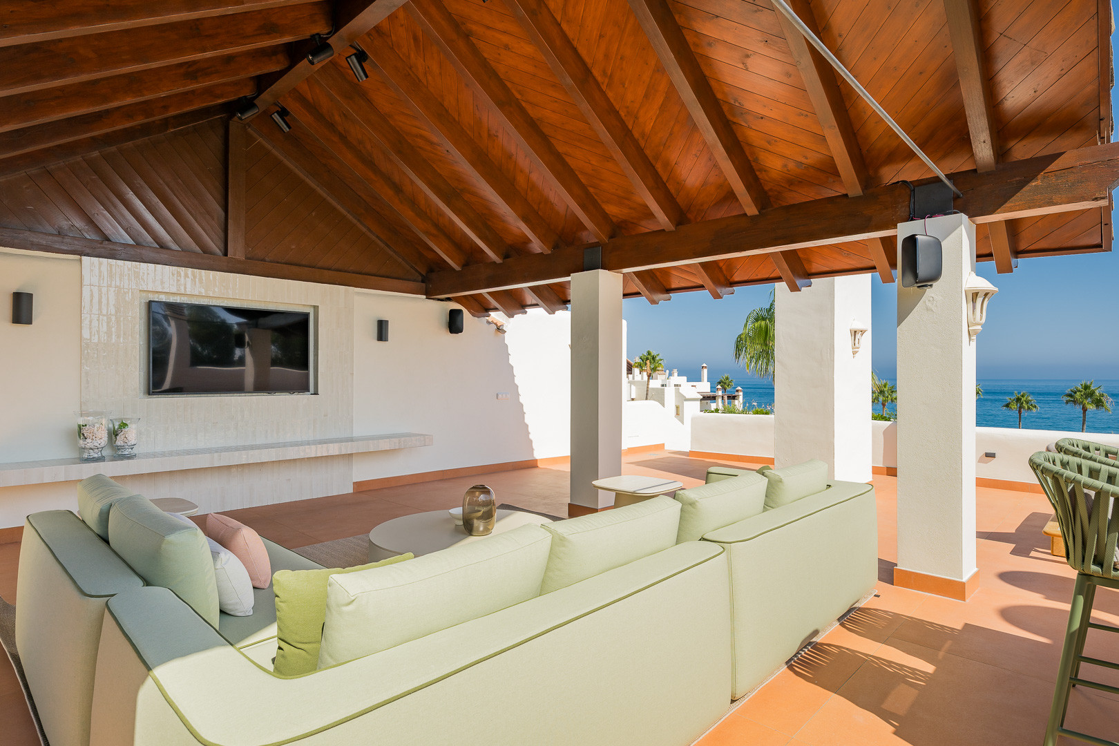 Covered outdoor seating area with a television, a large sofa, and a partial ocean view.
