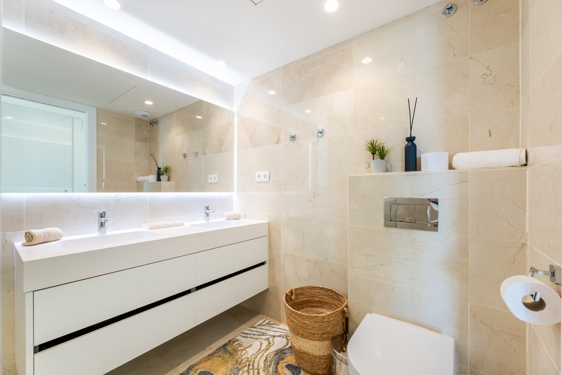 Modern bathroom with dual sinks, marble walls, and decorative plants on a shelf.