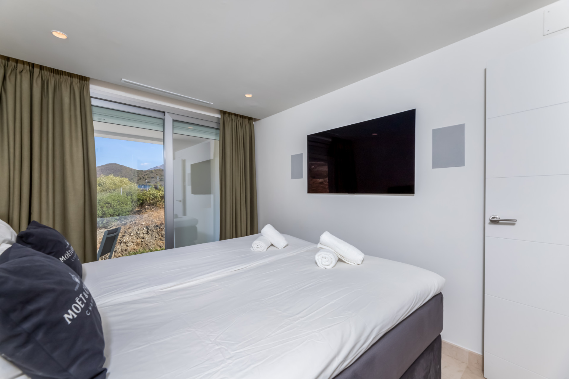 Bedroom view from a different angle featuring a wall-mounted TV and large windows with curtains.