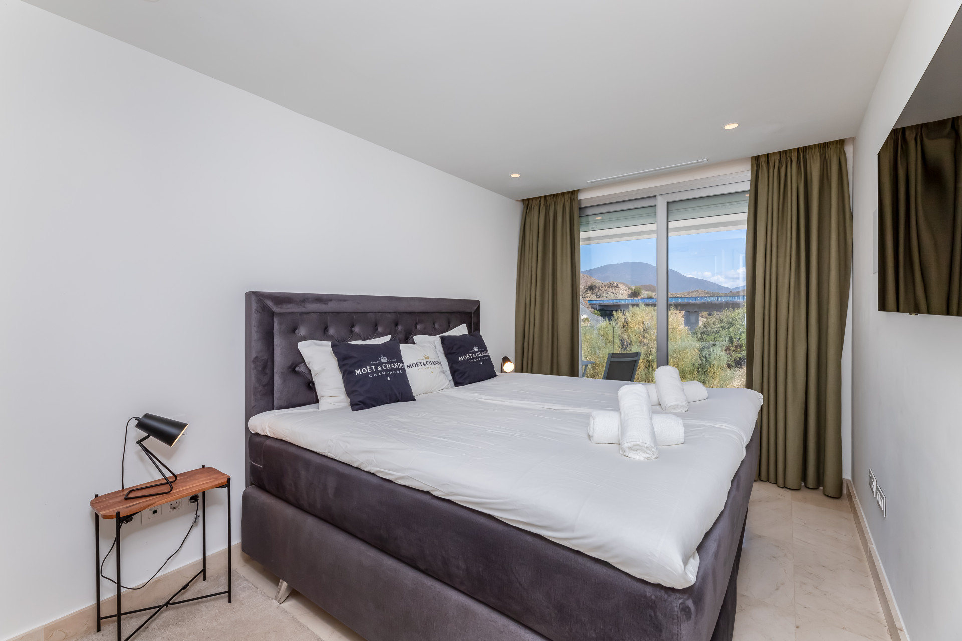 Bright bedroom with a large bed, modern decor, and curtained windows showcasing mountain views.