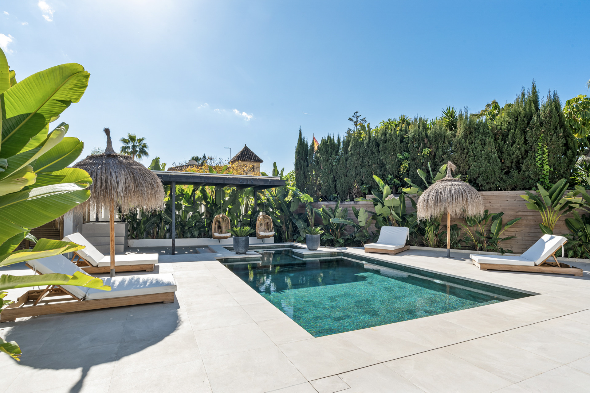 Swimming pool scene with sun loungers, palm trees, and a thatched umbrella.
