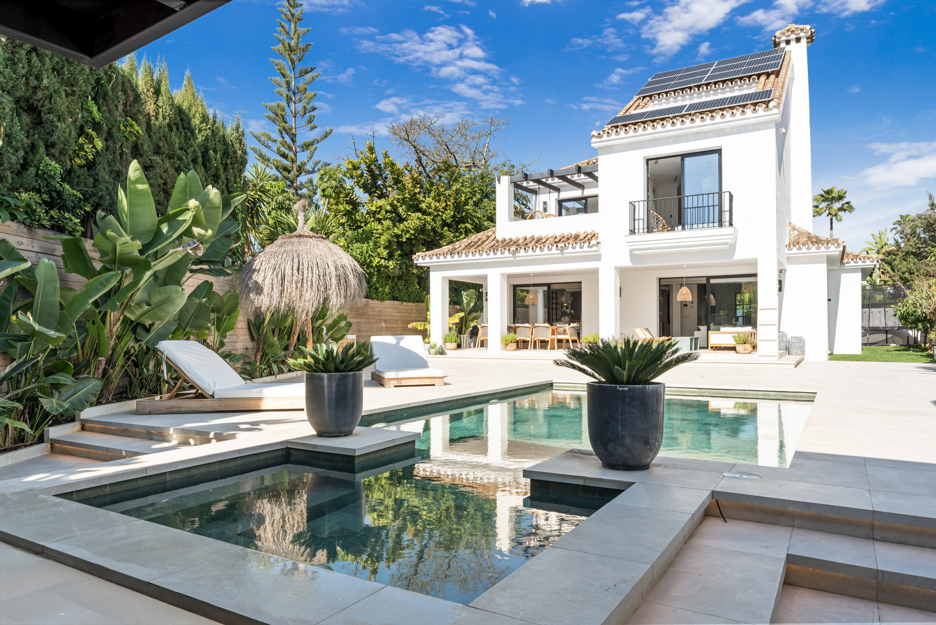 View of the pool area with loungers, tropical plants, and the villa in the background under a clear sky.