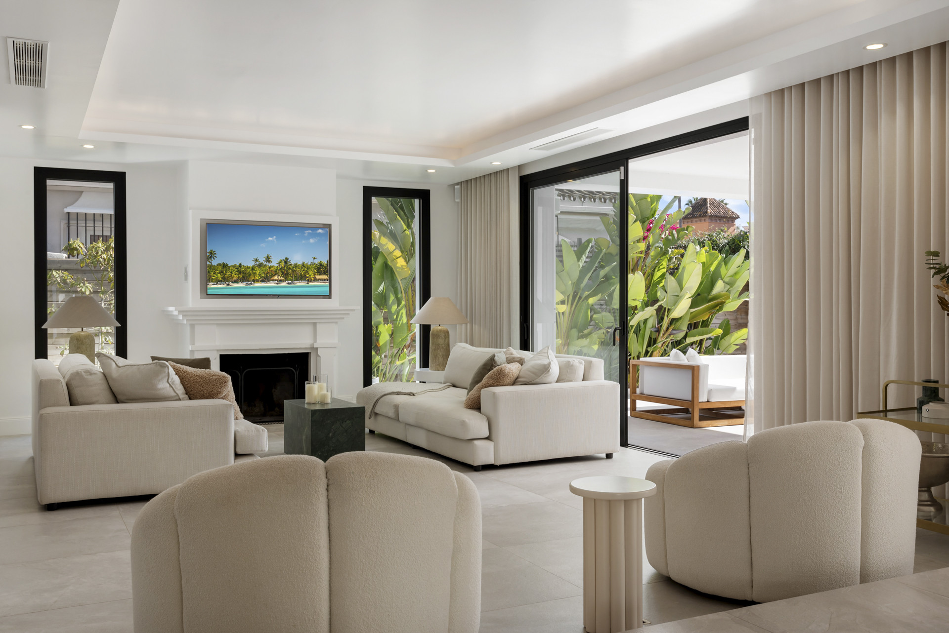 Living room with large windows, light-colored sofas, and a mounted TV above the fireplace.