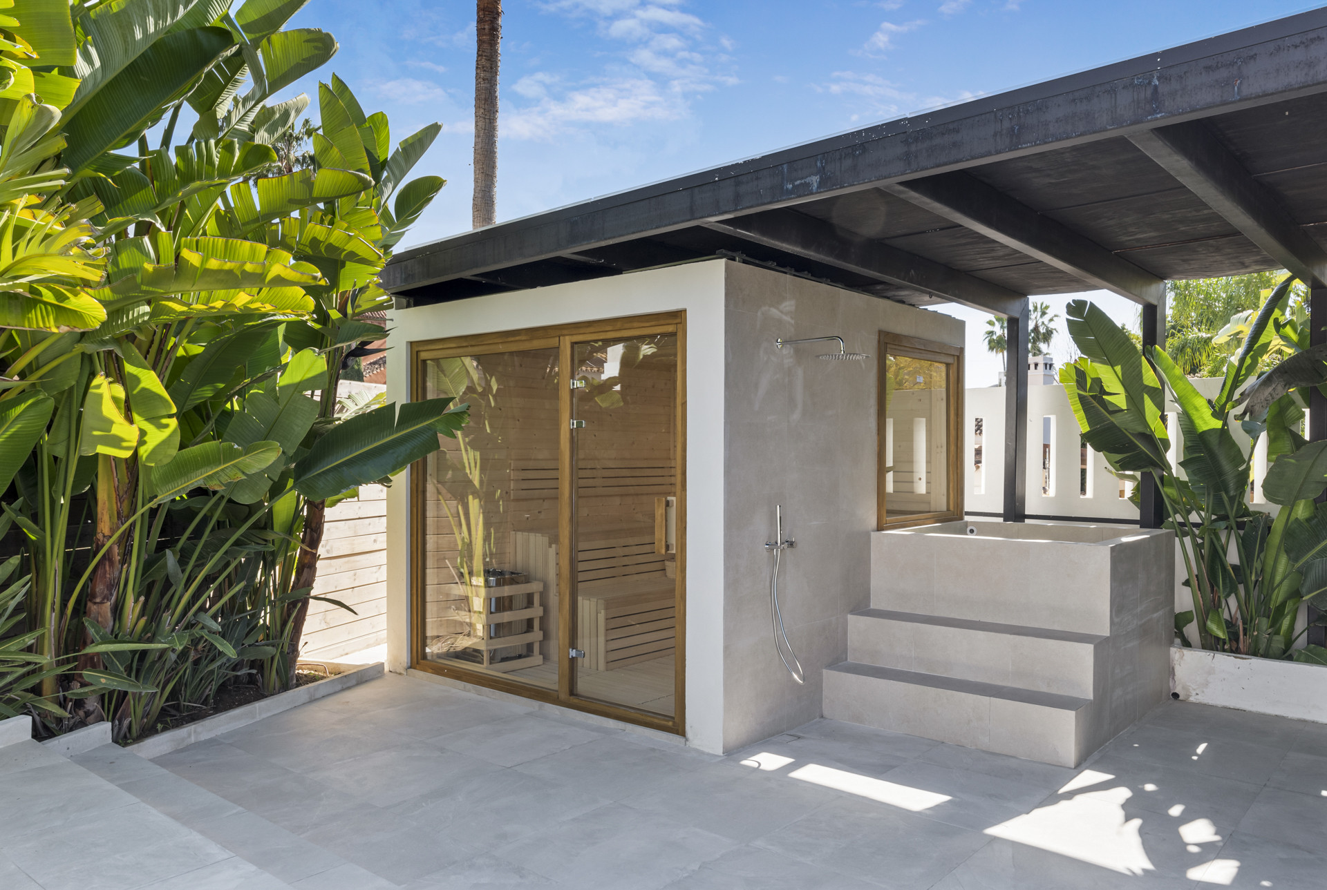 Outdoor sauna structure with wooden panels, glass doors, and an outdoor shower area surrounded by greenery.