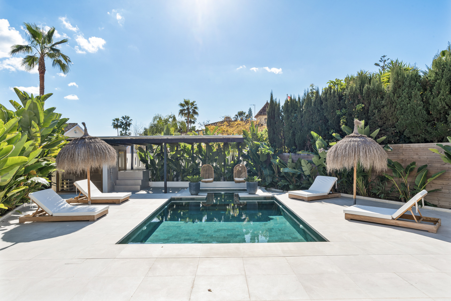 Clear view of the pool surrounded by loungers and tropical plants under a bright sky.