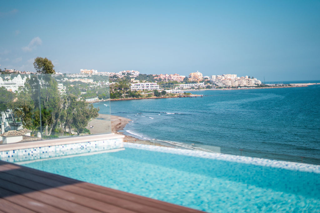 Infinity pool edge with wooden decking overlooking the sea and a distant coastline with buildings.