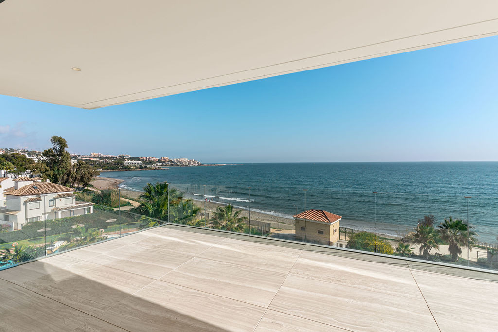 Balcony with light-colored tiled floor and glass railing overlooking a beach and sea.