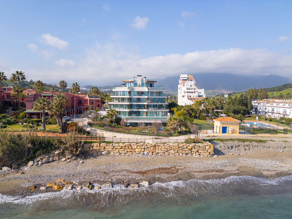 Aerial view of a modern beachfront building with surrounding residences, palm trees, and mountains.