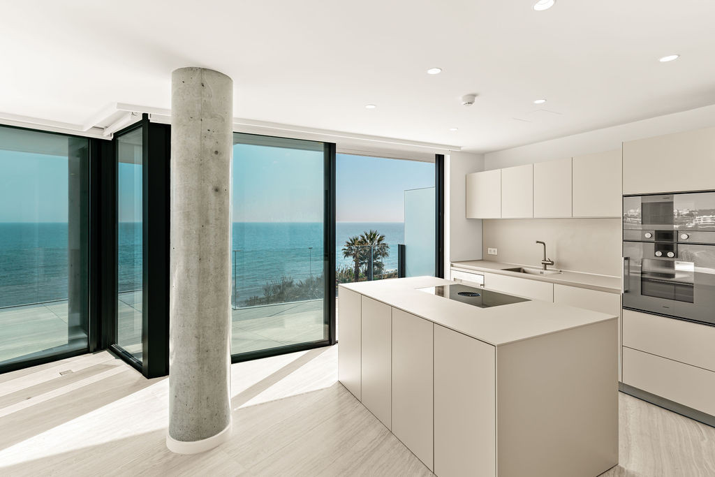 Wide shot of a kitchen with light grey cabinets and island, a concrete column, and floor-to-ceiling ocean views.