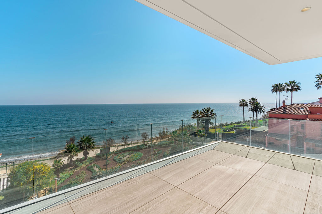 Outdoor terrace with light-colored tiles, glass railing, ocean view, and palm trees.
