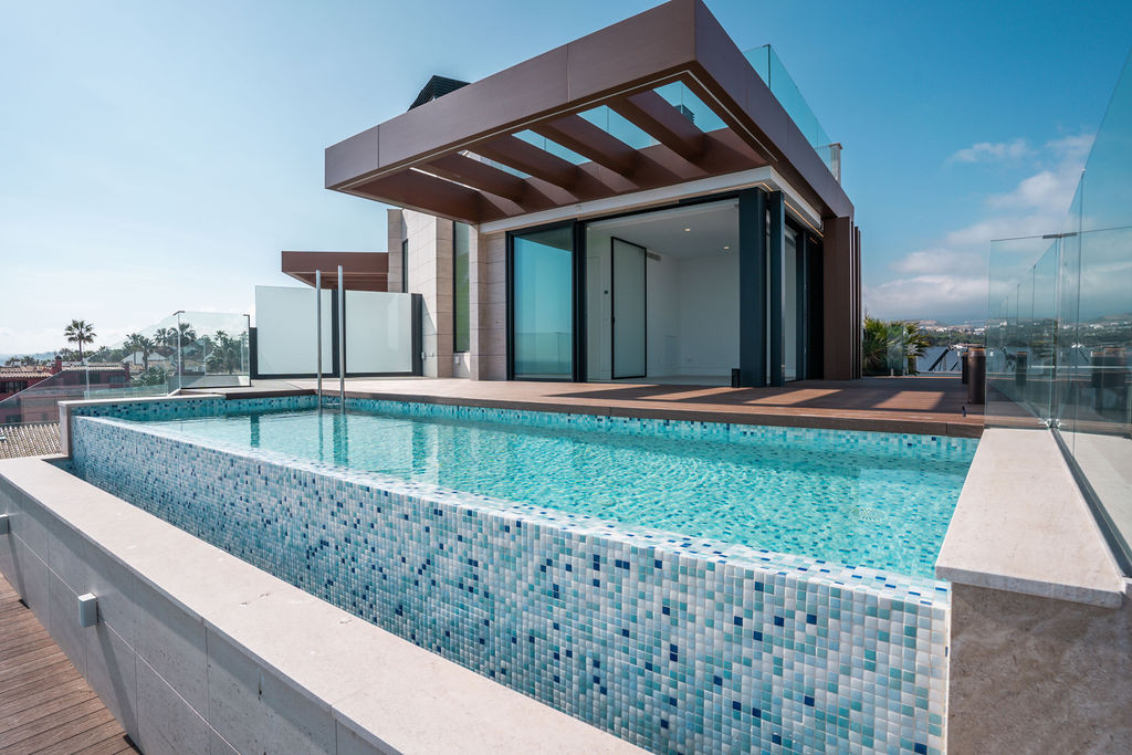 Rooftop infinity pool with mosaic tiles, wooden deck, and modern building with glass doors.