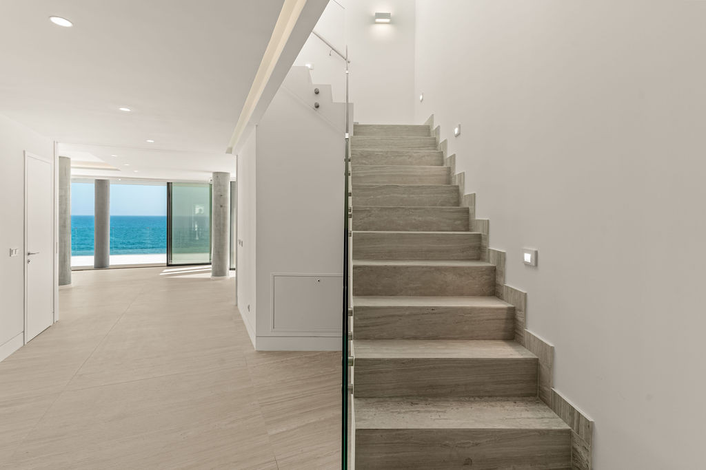 Interior view of a modern hallway with a staircase, leading to an ocean view through glass doors.