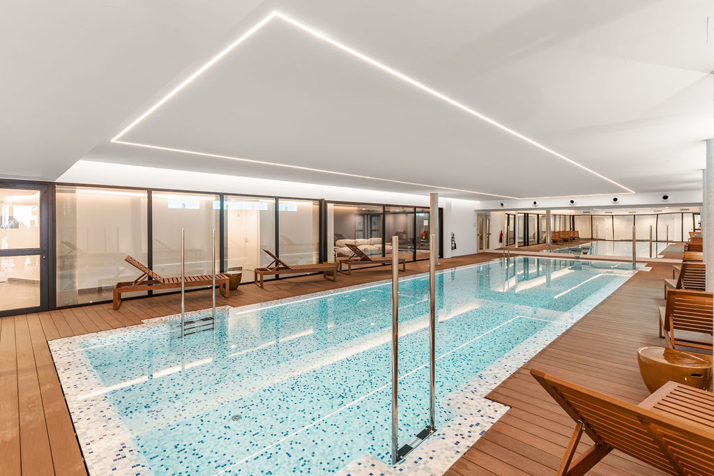Indoor swimming pool with light blue tiles, wooden decking, and lounge chairs.