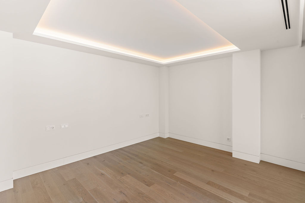 Corner of an empty room with light wood flooring, white walls, and a recessed ceiling light.