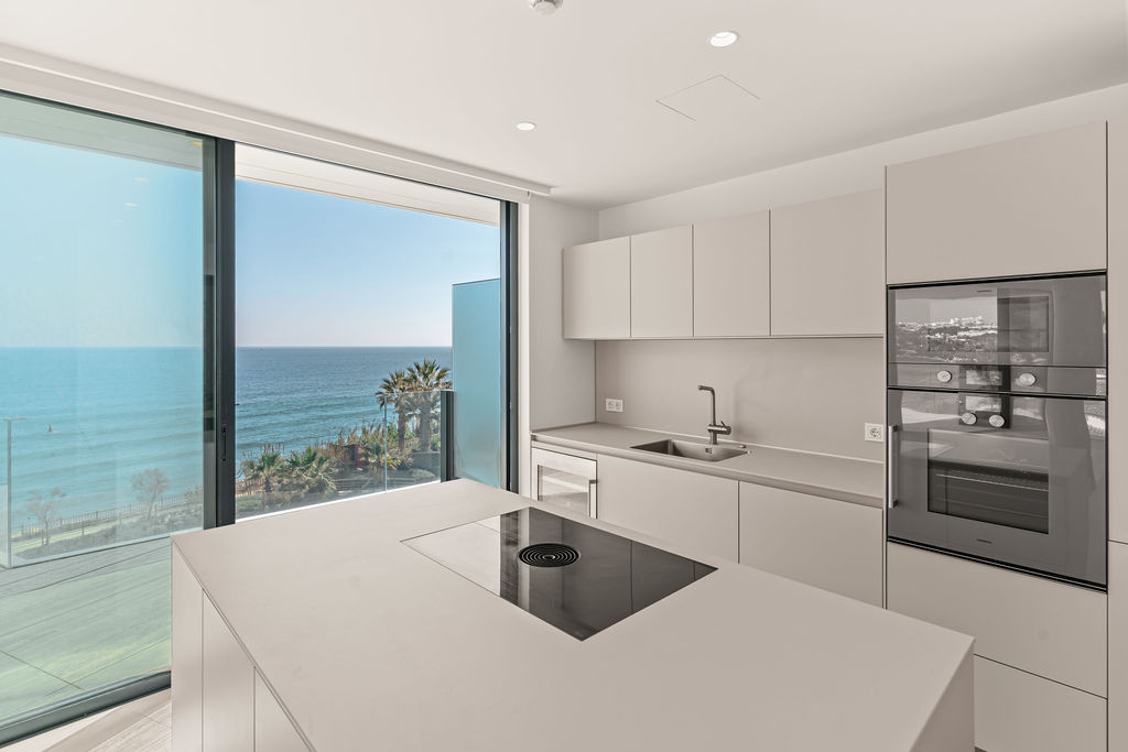 Kitchen with light grey island and built-in stovetop, overlooking the ocean and palm trees through a large window.