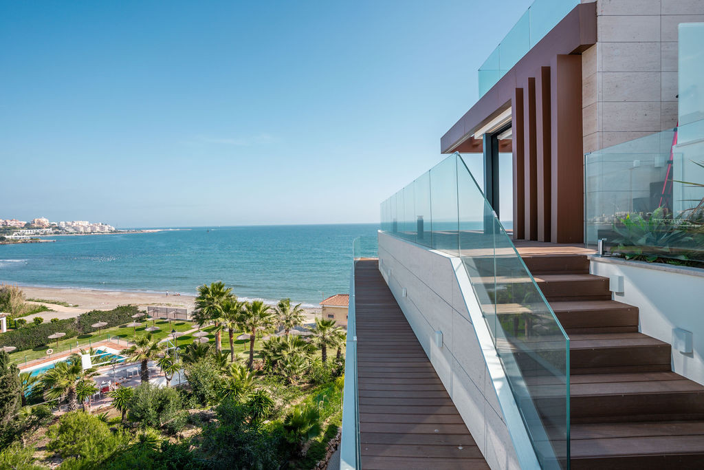 Wooden staircase with glass railing leads to an upper balcony, overlooking a beach and town.