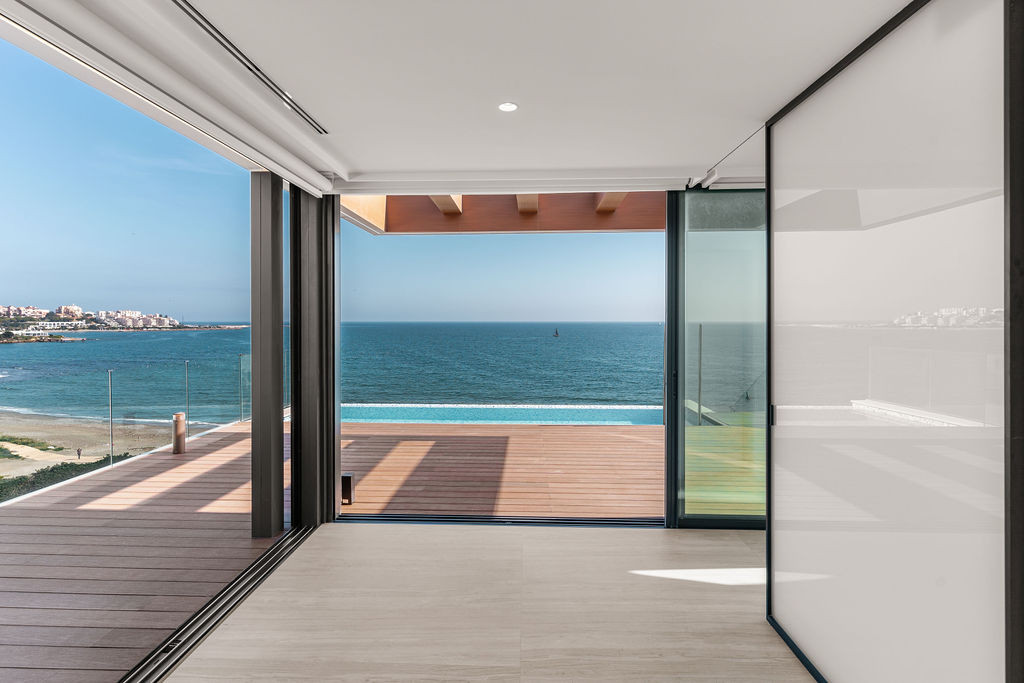 Indoor room with beige floor, open glass doors, leading to an outdoor deck and sea view.