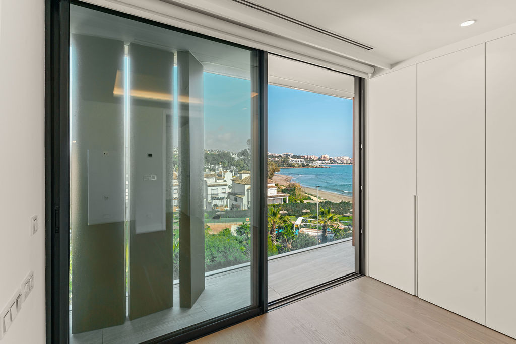 Bedroom with sliding glass doors opening to a balcony, revealing a beach, pool, and town.