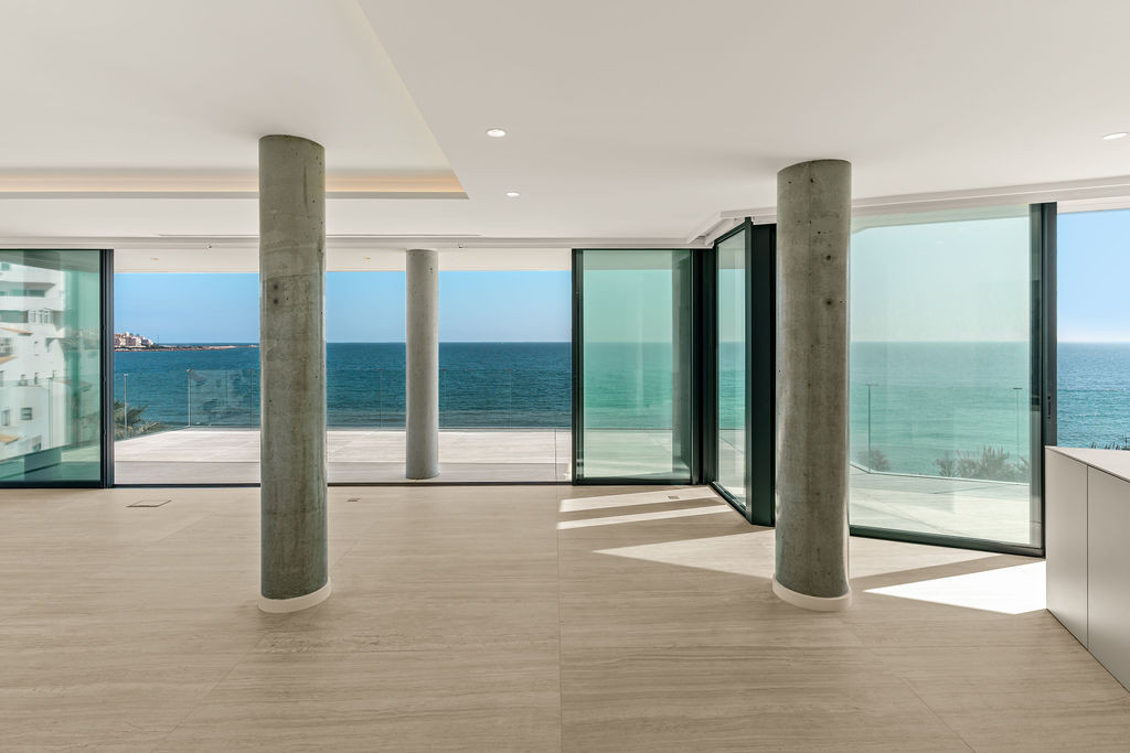 Spacious empty room with concrete columns, light tiled floors, and expansive sliding glass doors facing the ocean.