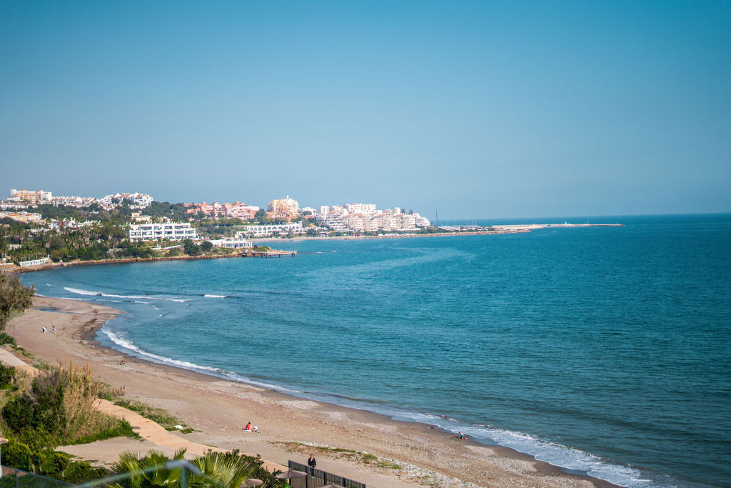 Coastal view of a sandy beach, blue ocean, and a distant town on a sunny day.