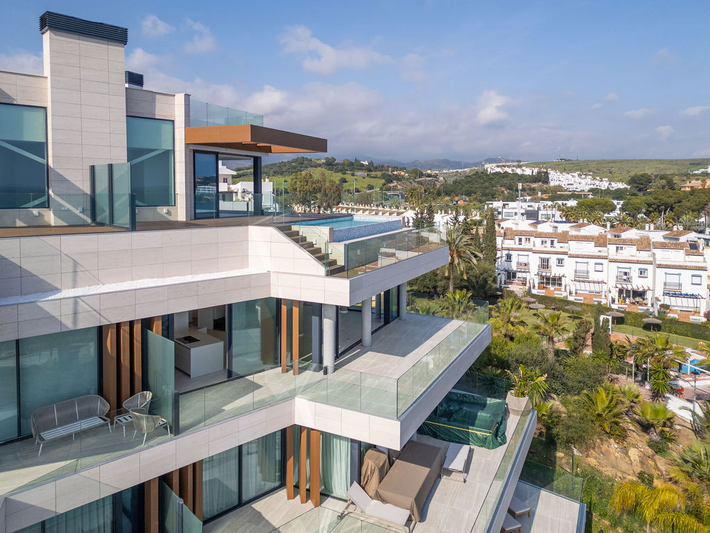 Exterior shot of a terraced building with multiple balconies, a pool, and distant town views.