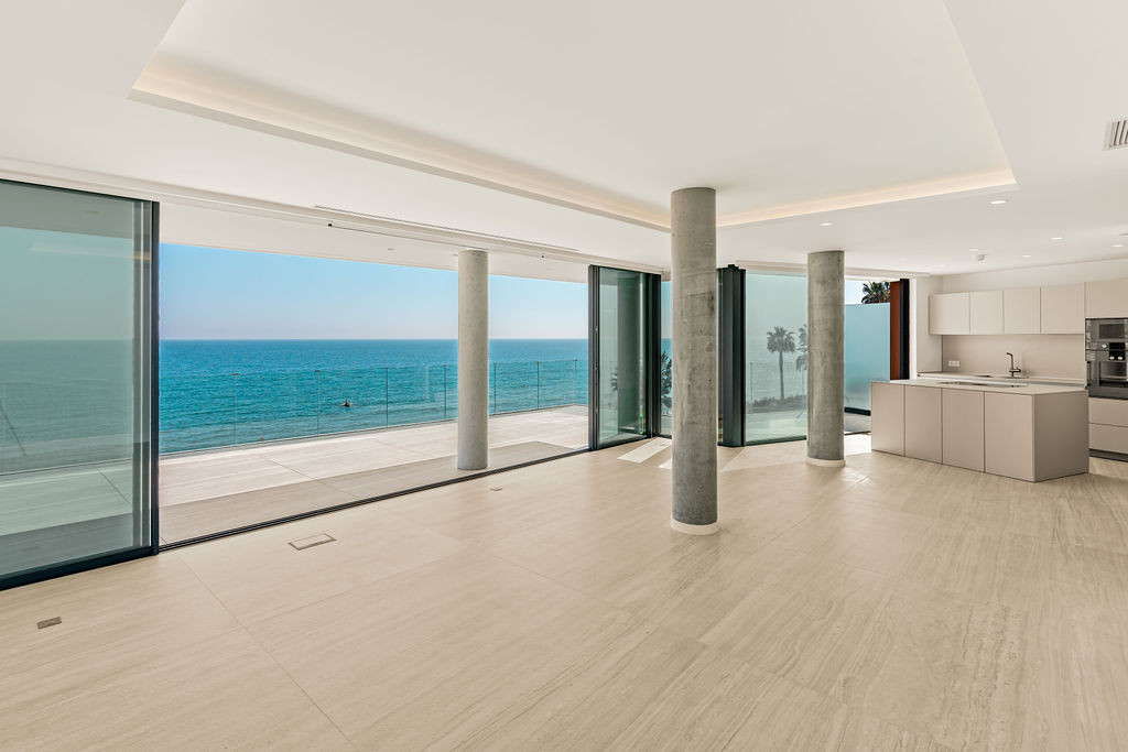 Large room with concrete columns, expansive ocean views, and a modern kitchen area visible.