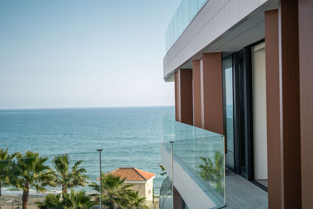 View from a balcony with a glass railing towards the ocean and palm trees under a clear sky.