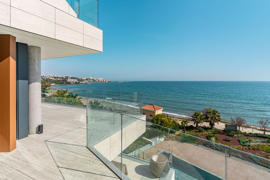 Outdoor terrace with glass railing overlooks the sea and distant coastline under a clear sky.