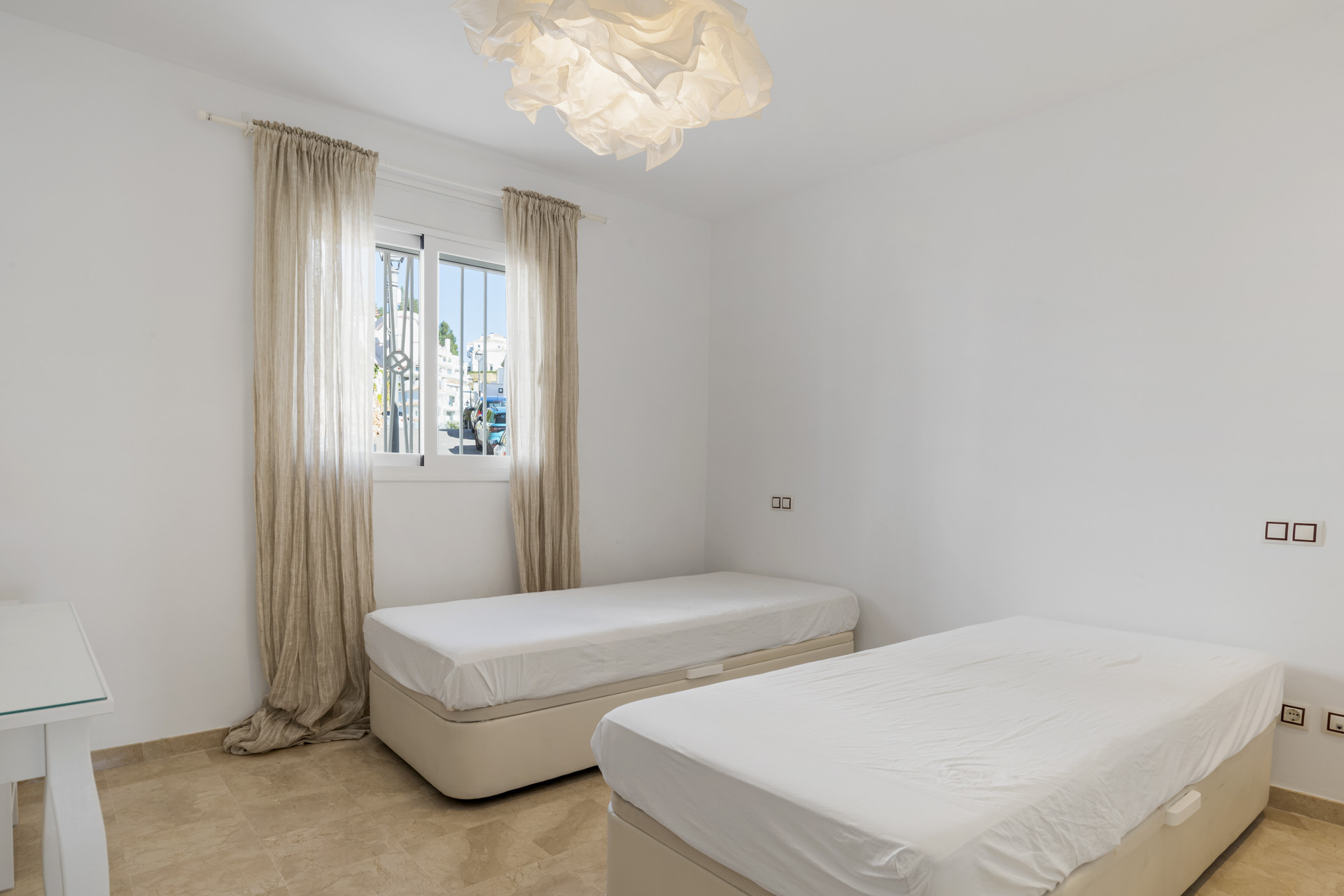 Bedroom with two beds, a window with beige curtains, and an exterior view of buildings and trees.