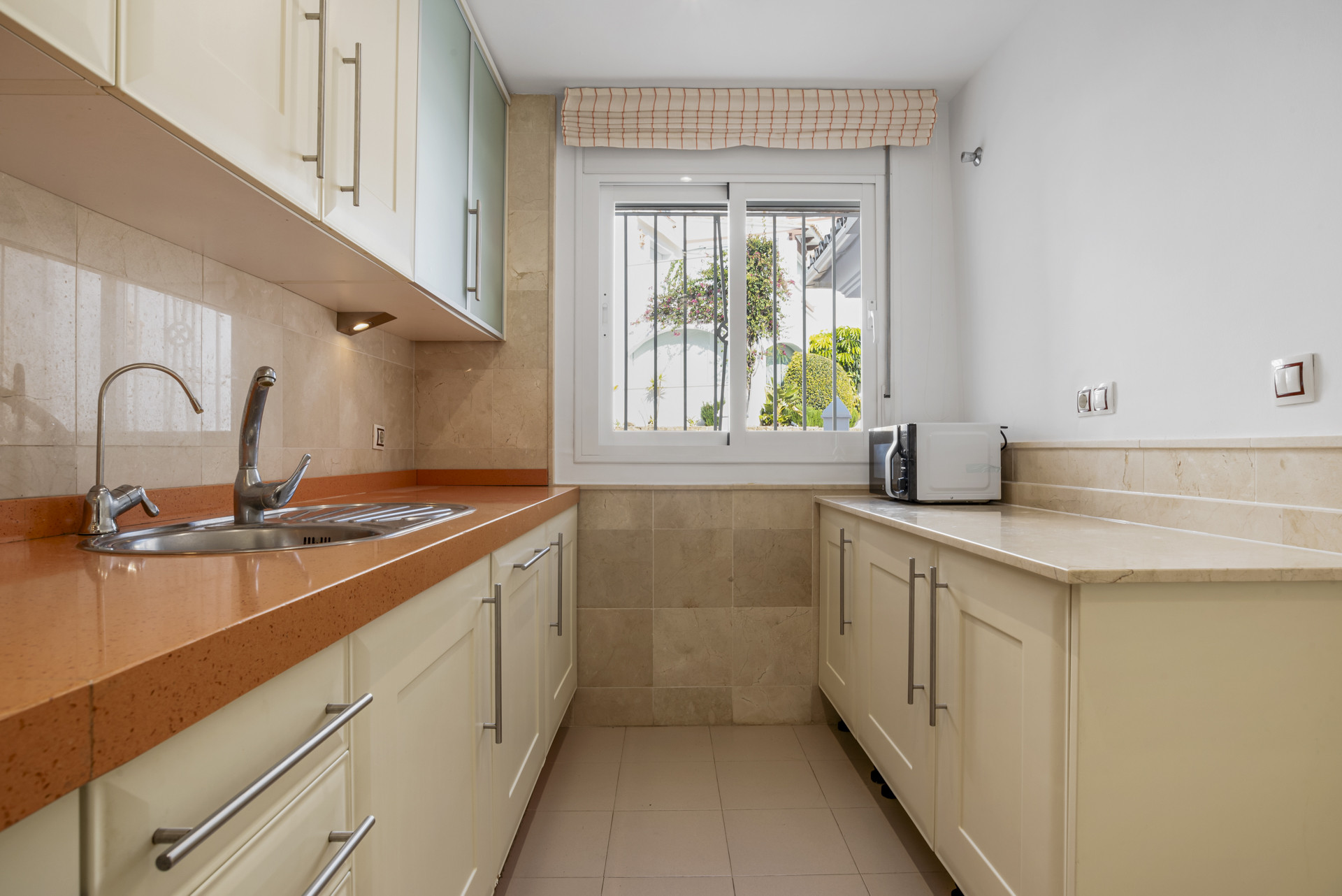 Galley kitchen showing light cream cabinets, a sink, and a window with white blinds and security bars.