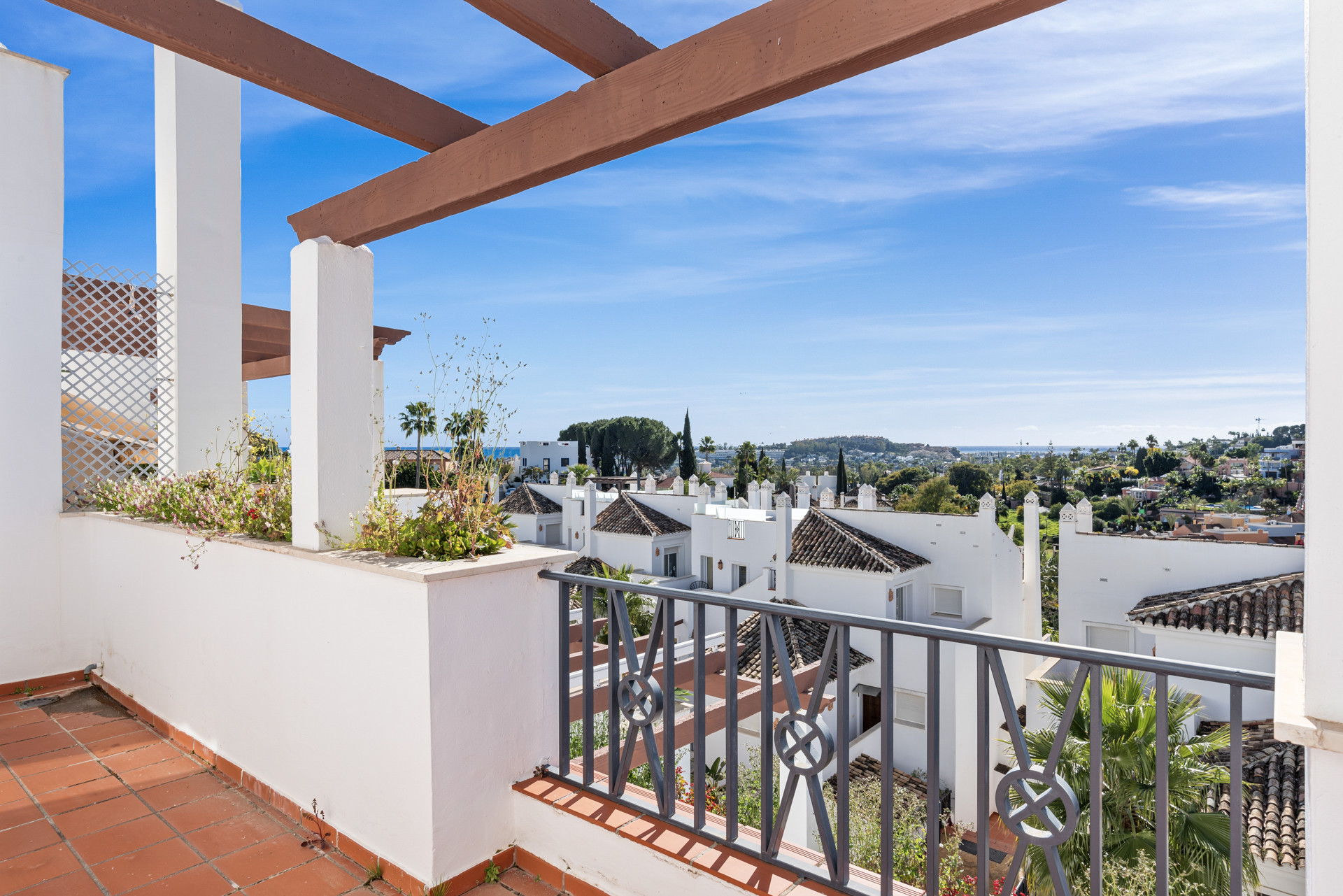 Balcony with terracotta tiles, white railing, wooden pergola beams, overlooking a town and hills.