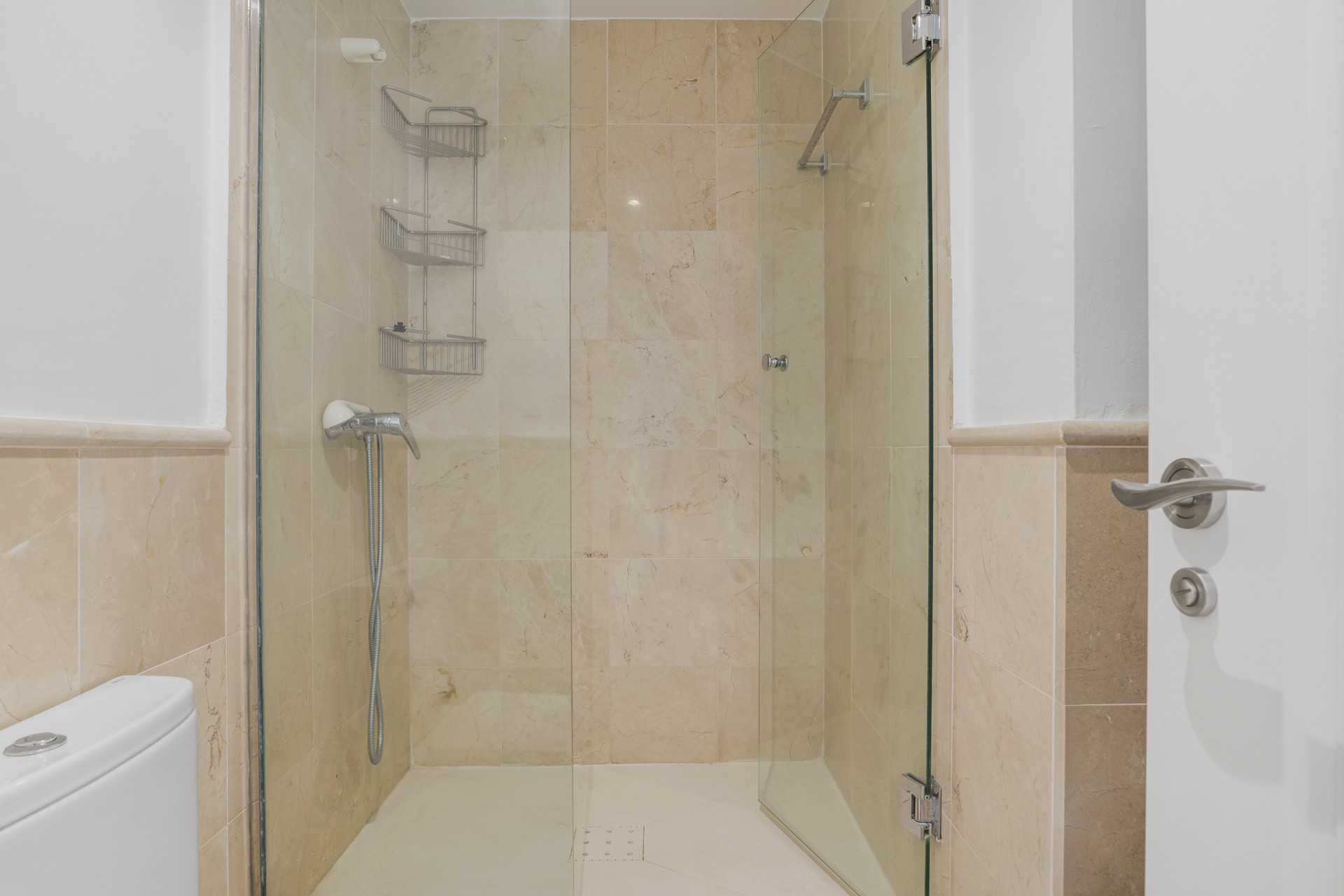 Walk-in shower with clear glass screen, light beige tiles, a chrome showerhead, and storage rack.