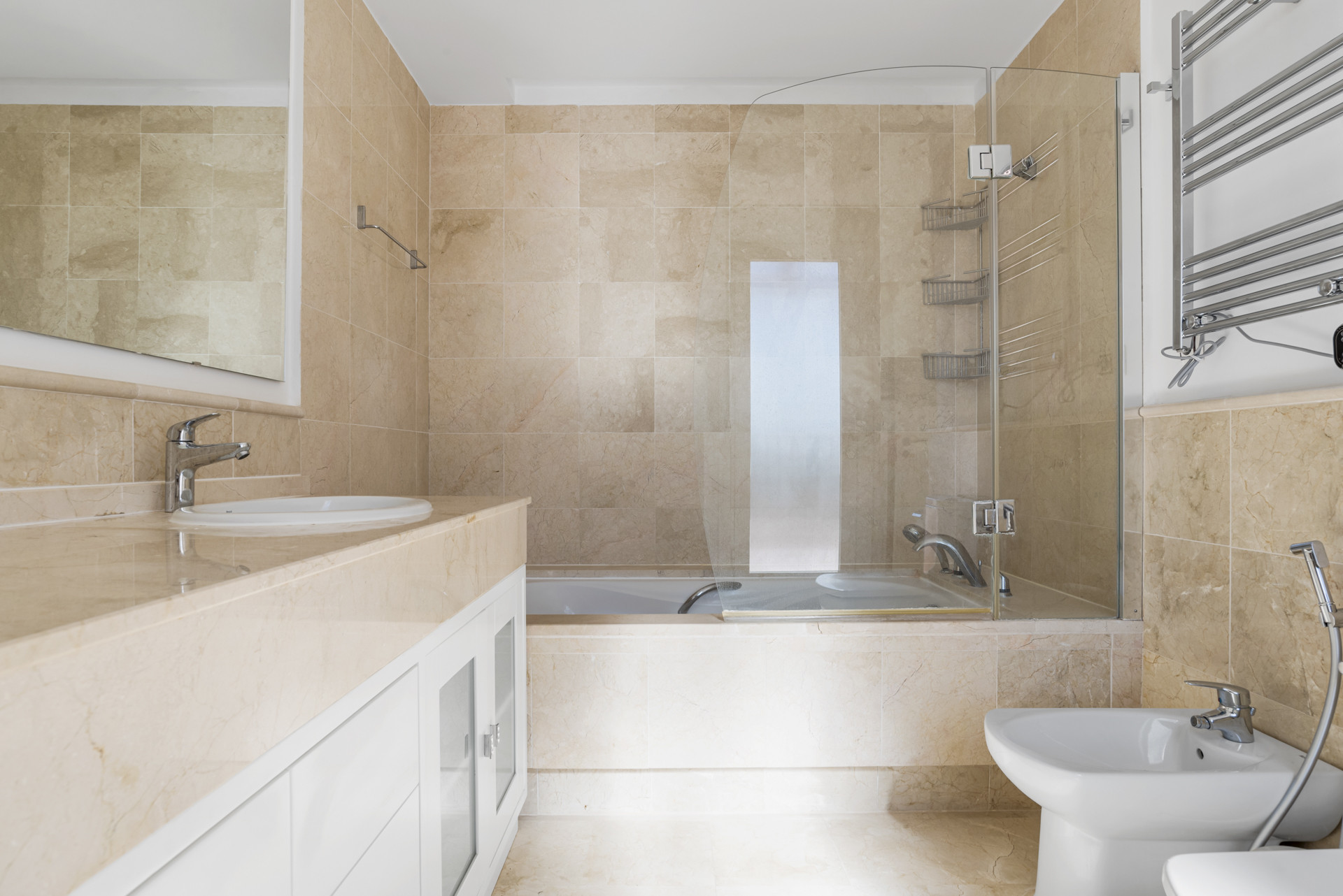 Bathroom featuring light beige tiled walls and floor, a bathtub with glass screen, sink, and bidet.
