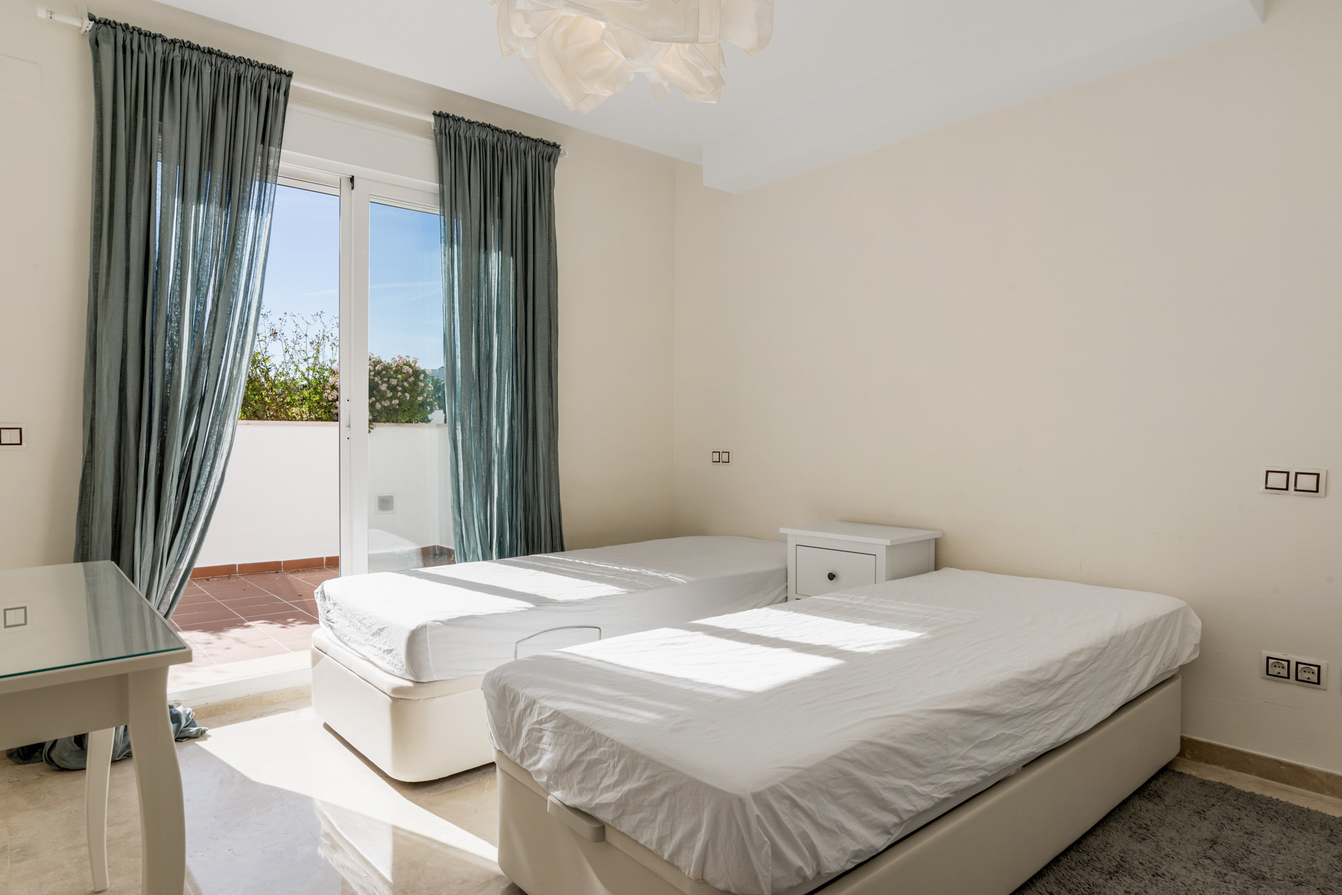 Bedroom with two beds, a glass table, and a balcony with blue curtains and distant greenery.