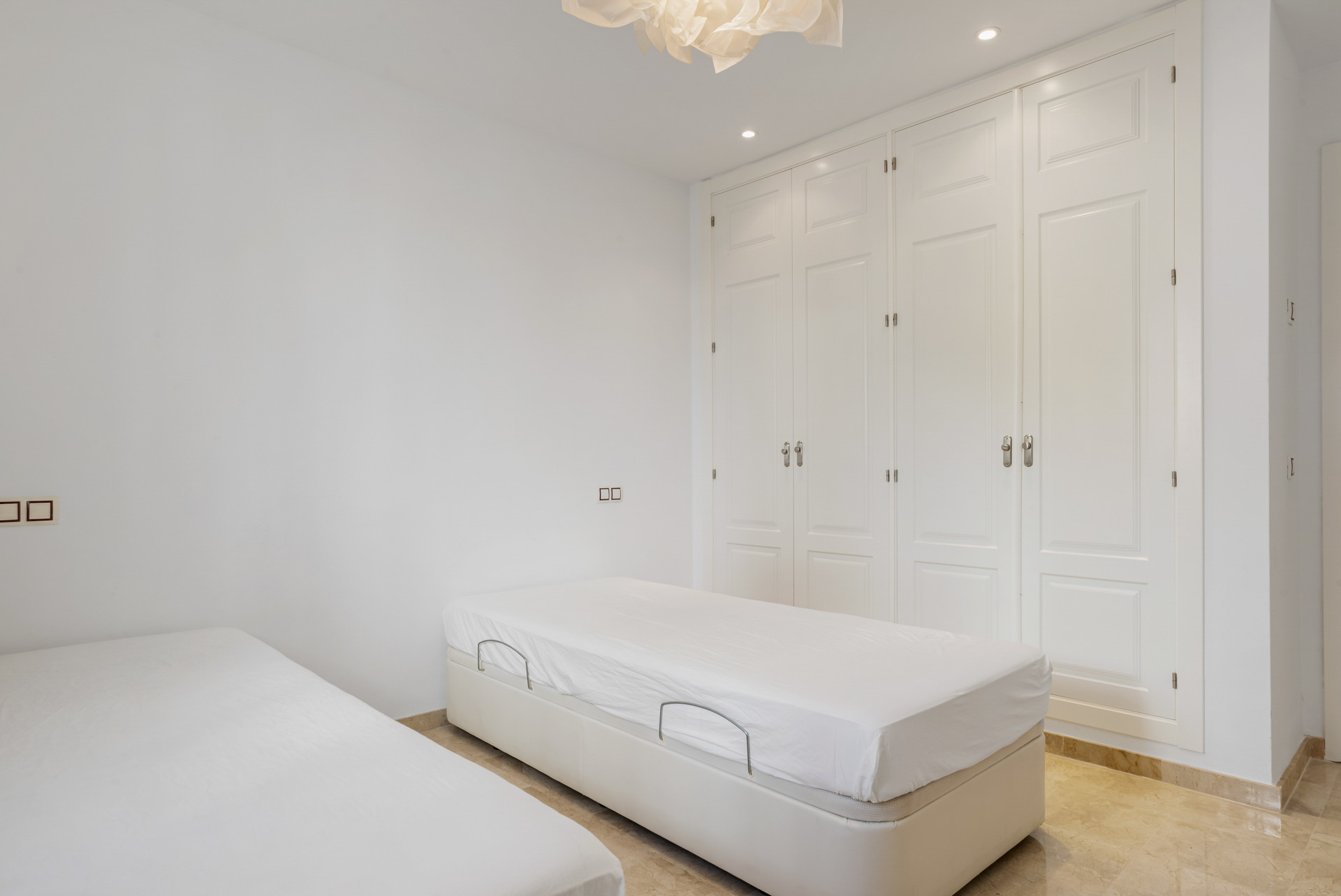 Bedroom with two twin beds and a wall of white built-in wardrobes, with a beige tiled floor.
