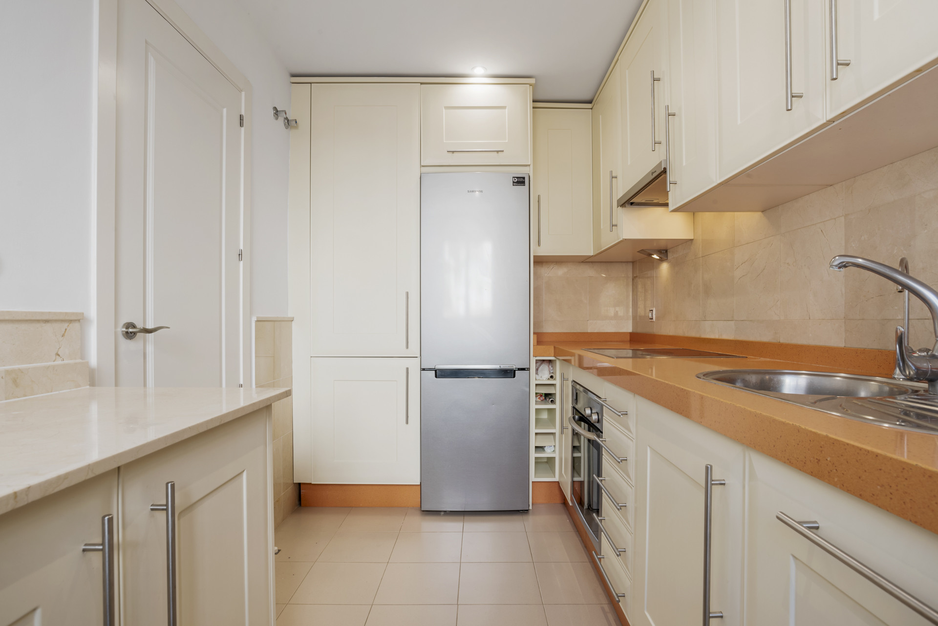 Galley kitchen with cream cabinets, stainless steel refrigerator, and light orange countertops.