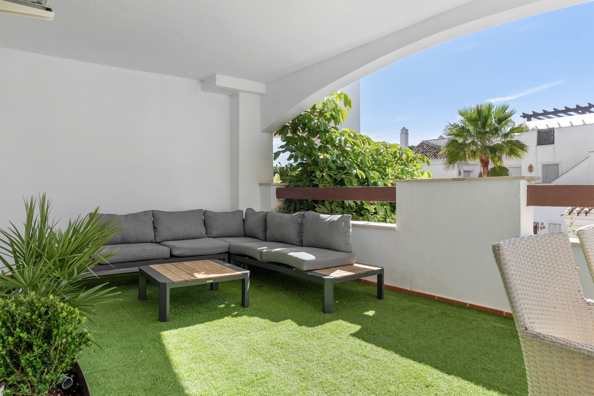Cozy terrace with gray sectional sofa and a small table, surrounded by greenery and clear sky.