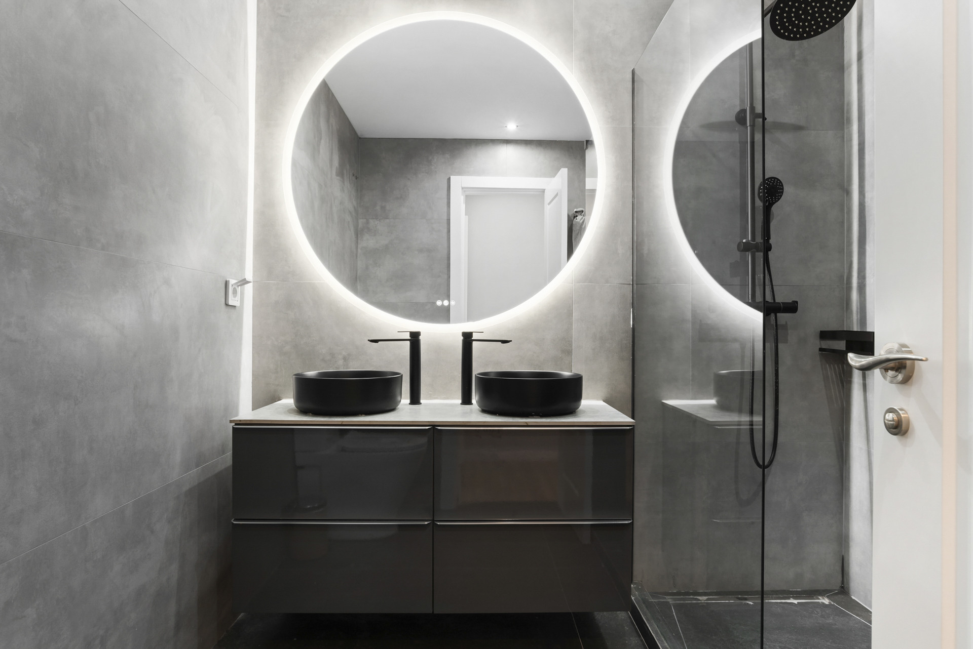 Modern bathroom with black sinks, a circular illuminated mirror, and a glass shower enclosure.