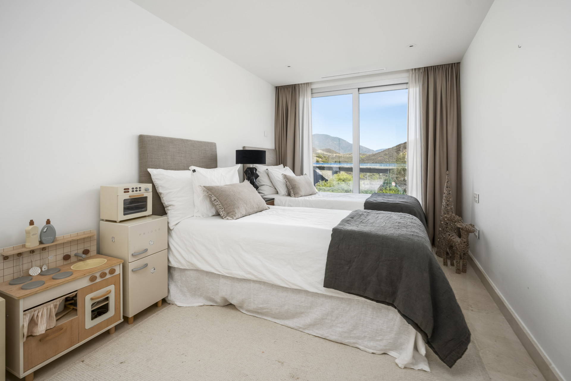 Bright bedroom with twin beds, large windows, and a children's play kitchen set.