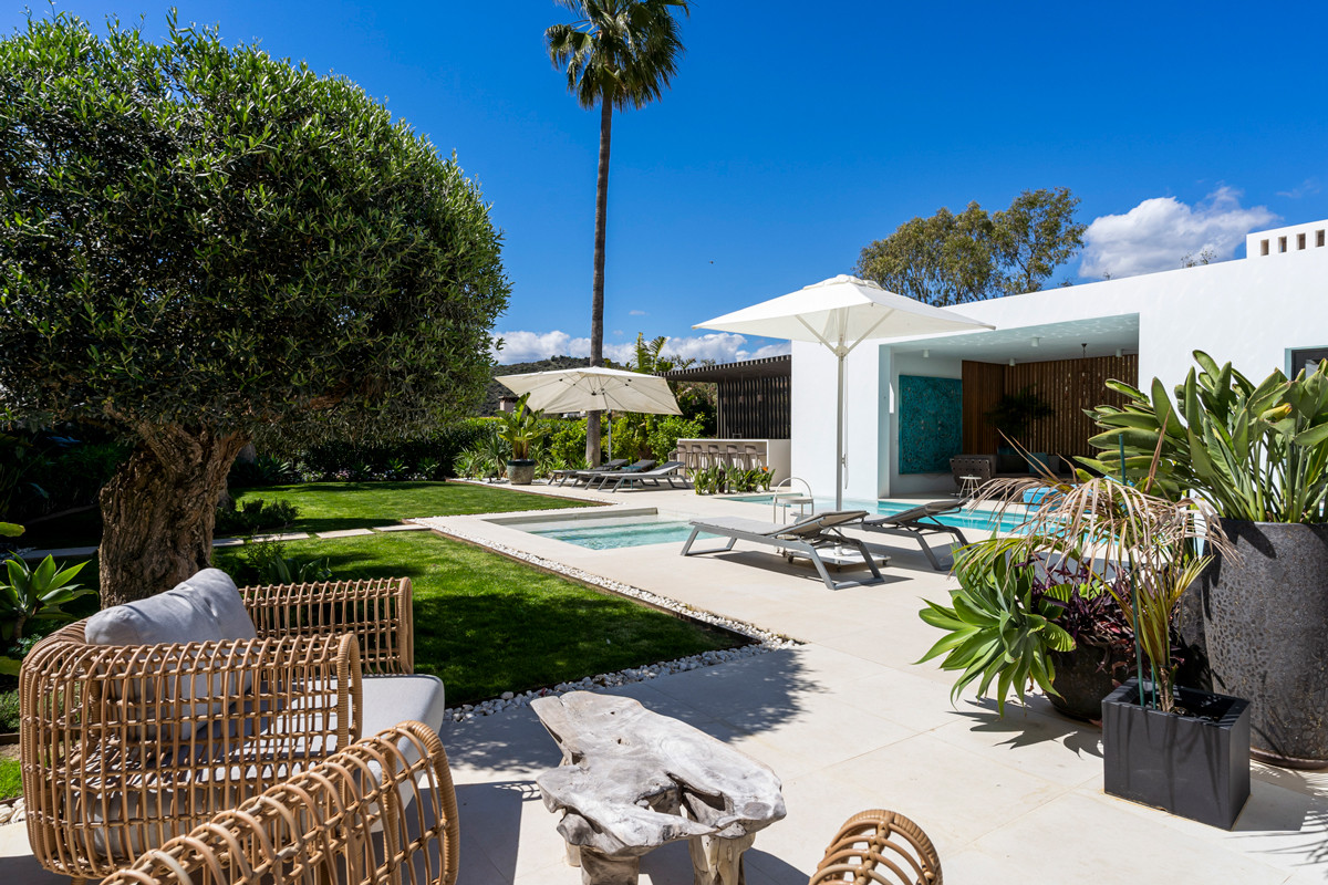 Outdoor lounge chairs and a wooden table on a tiled patio, surrounded by a lawn and a swimming pool.