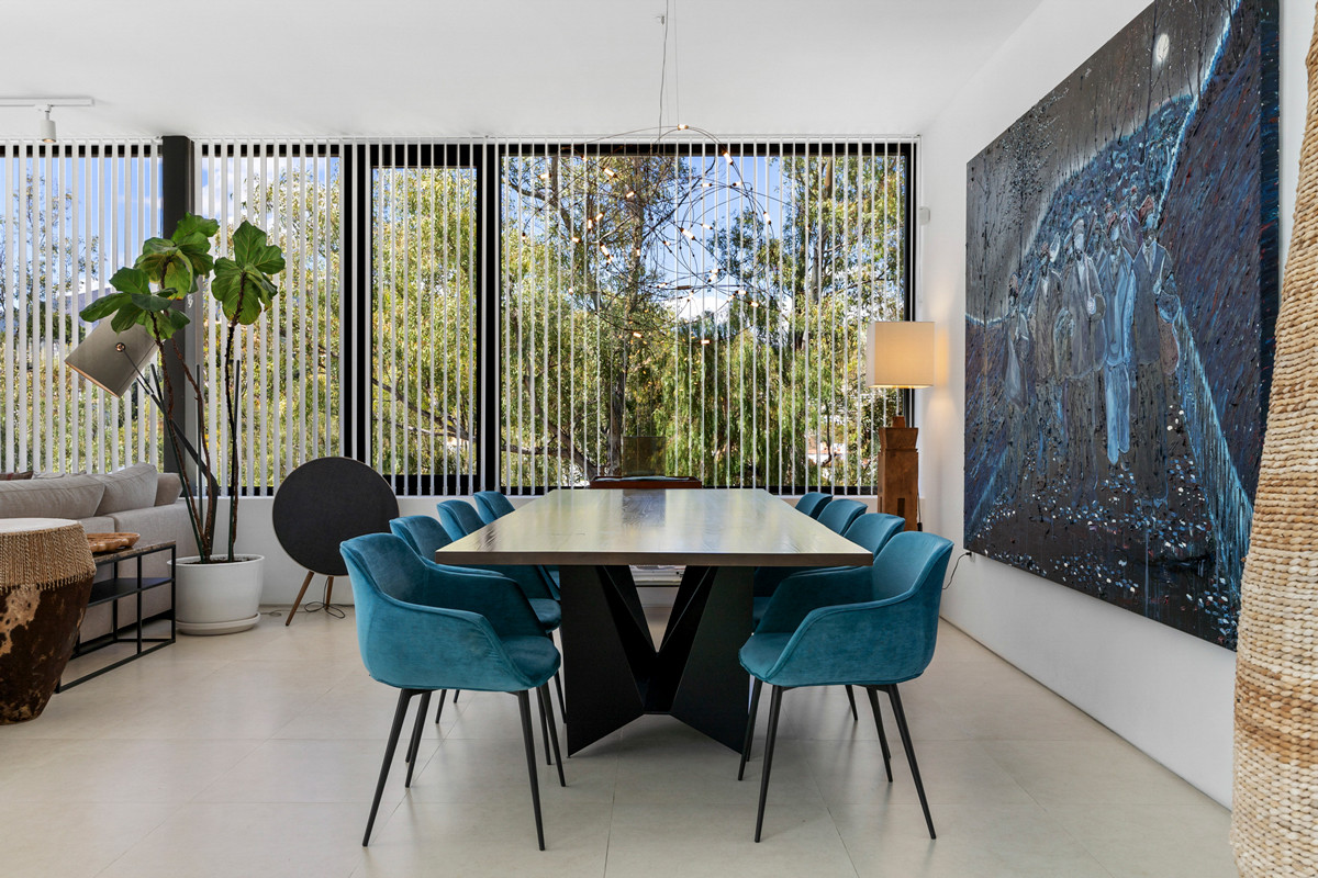 Dining area with large table, teal chairs, and abstract art, facing tall windows with outdoor views.