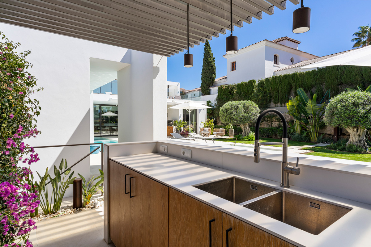 Outdoor kitchen counter with two sinks and a faucet, overlooking a patio, garden, and another villa.