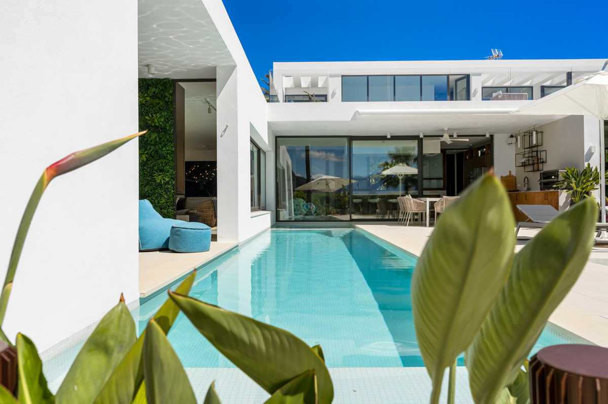 View from a lush garden towards a narrow pool, an outdoor seating area, and the villa's glass facade.