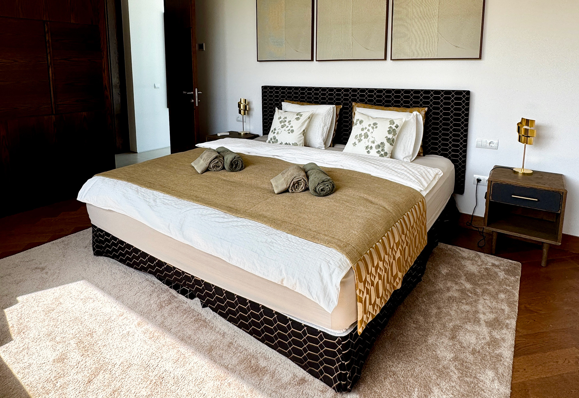 Bedroom with a large bed, patterned headboard, nightstands, gold lamps, and artwork above the bed.