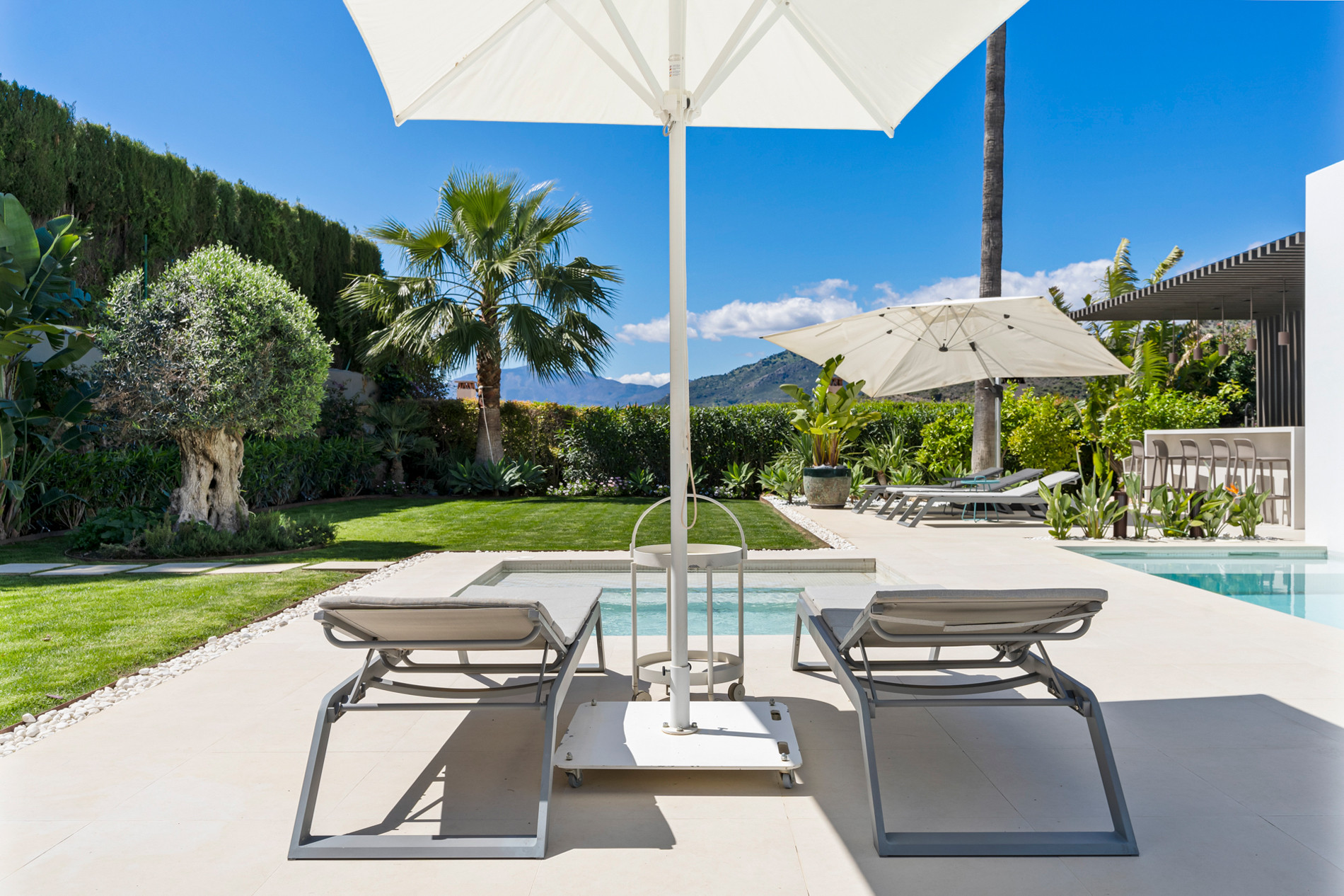 Two grey sun loungers under a white umbrella, with a lush green lawn and mountains in the background.