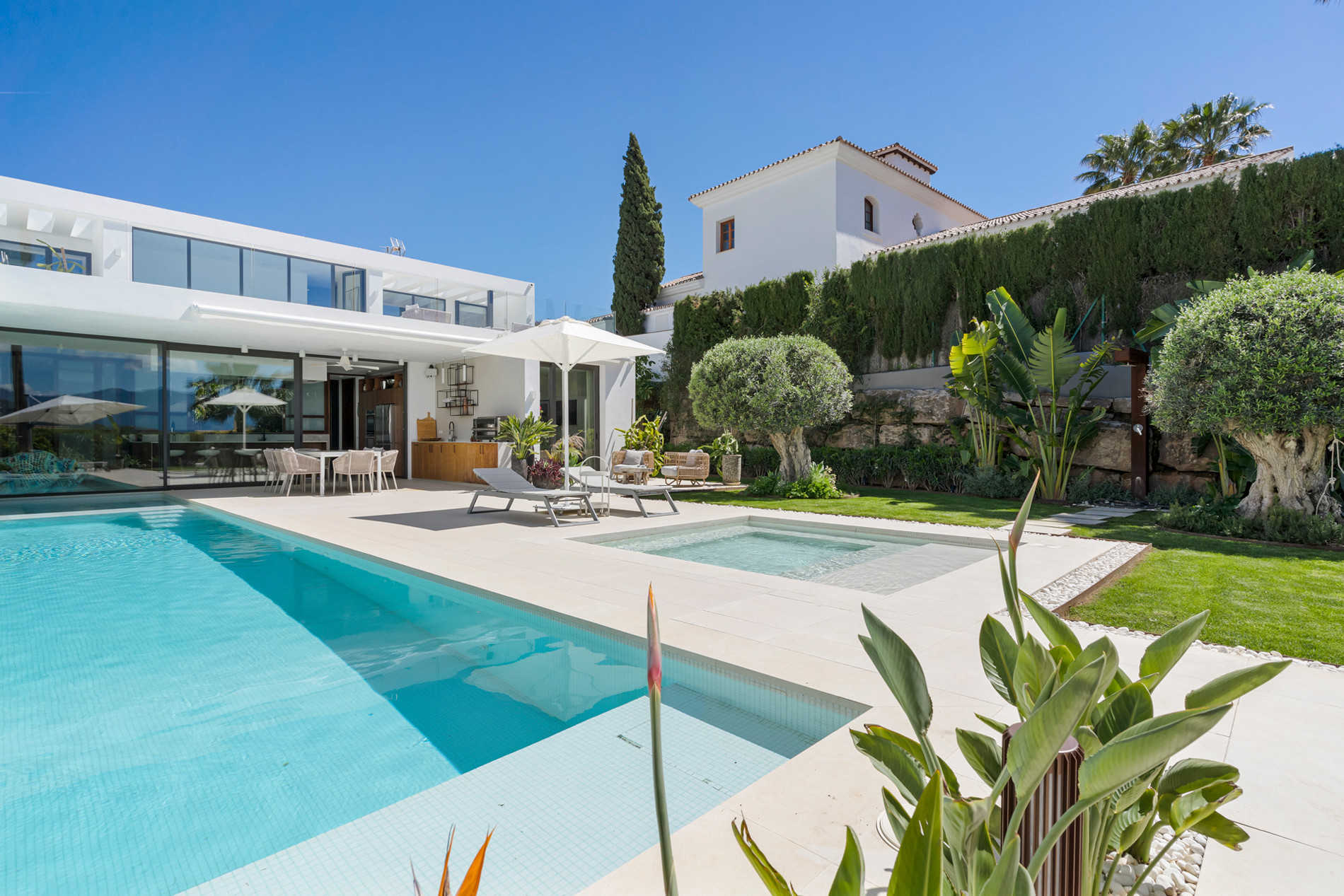 Exterior view of a modern white villa with a large pool, a smaller hot tub, and a landscaped garden.