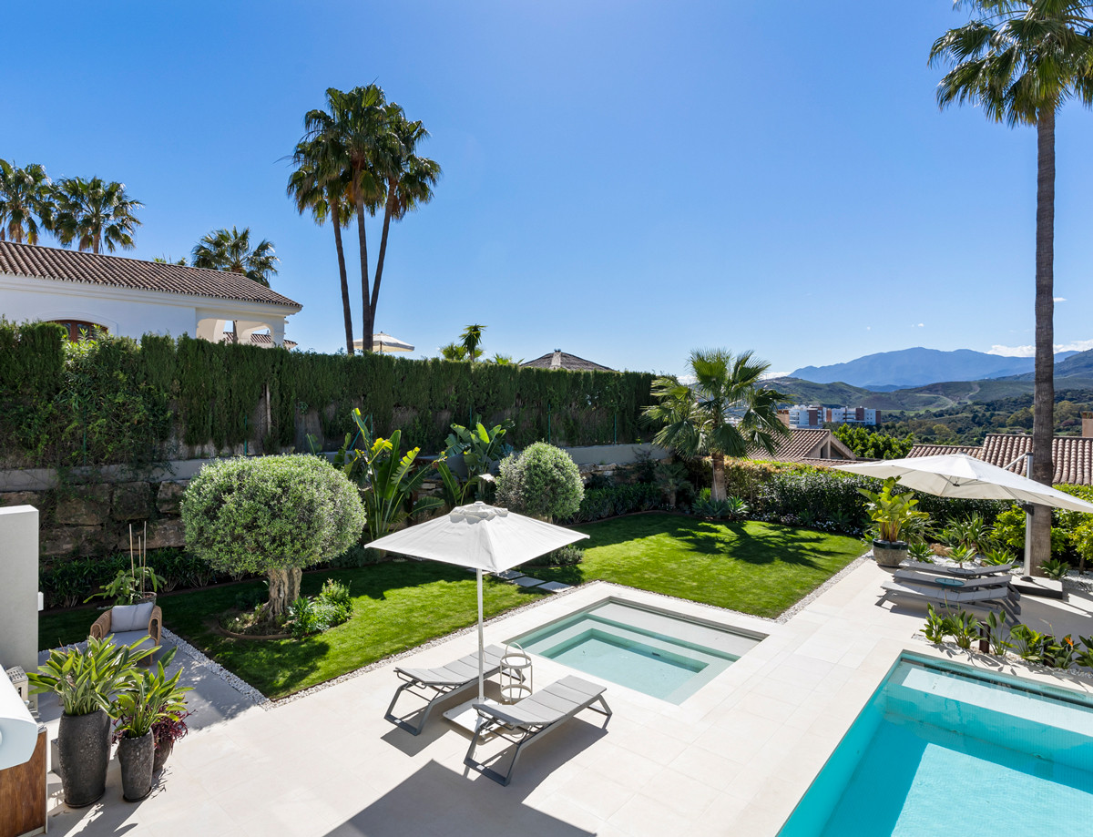 Aerial view of a luxury villa's outdoor area with two pools, a manicured lawn, and distant mountains under a blue sky.